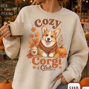 May include: A sand-colored crewneck sweatshirt featuring a graphic print of a Corgi wearing a brown scarf, surrounded by autumn leaves, a pumpkin, and a mug with a heart. The text reads "cozy Corgi Club" in a brown, retro-style font.