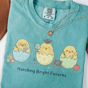 Puede incluir: Una camiseta turquesa Comfort Colors con tres pollitos de dibujos animados saliendo de huevos, cada uno con objetos escolares. La camiseta tiene el texto "Hatching Bright Futures". Un collar plateado est&aacute; sobre la camiseta.