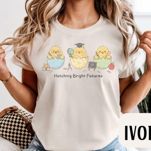 Puede incluir: Camiseta de color marfil con un dise&ntilde;o de tres pollitos de dibujos animados saliendo de huevos, cada uno con objetos escolares. El texto "Hatching Bright Futures" est&aacute; impreso debajo. La parte superior es de cuello redondo y tiene mangas cortas.