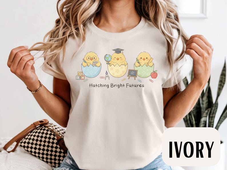 Puede incluir: Camiseta de color marfil con un dise&ntilde;o de tres pollitos de dibujos animados saliendo de huevos, cada uno con objetos escolares. El texto "Hatching Bright Futures" est&aacute; impreso debajo. La parte superior es de cuello redondo y tiene mangas cortas.