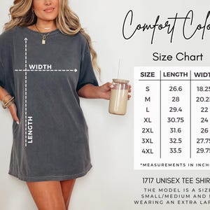 Puede incluir: Una camiseta unisex gris oscuro con una tabla de tallas que muestra las medidas en pulgadas. La camiseta tiene el texto "Comfort Colors" y "1717 Unisex Tee Shirt". La modelo lleva una talla extra grande.