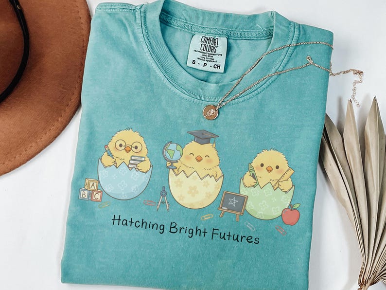 Puede incluir: Una camiseta turquesa Comfort Colors con tres pollitos de dibujos animados saliendo de huevos, cada uno con objetos escolares. La camiseta tiene el texto "Hatching Bright Futures". Un collar plateado est&aacute; sobre la camiseta.