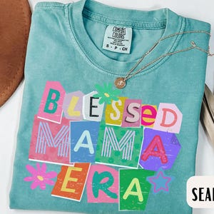 Peut inclure: T-shirt vert d'eau avec l'inscription "BLESSED MAMA ERA" en lettres colorées de style découpé. Le t-shirt a un col rond et des manches courtes. Un collier en or avec un pendentif repose sur le t-shirt. La chemise est de la marque Comfort Colors.