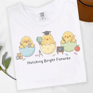 Puede incluir: Camiseta blanca con un dise&ntilde;o de tres pollitos de dibujos animados saliendo de huevos, cada uno con accesorios escolares. El texto "Hatching Bright Futures" est&aacute; impreso debajo. La camiseta tiene una etiqueta Comfort Colors.