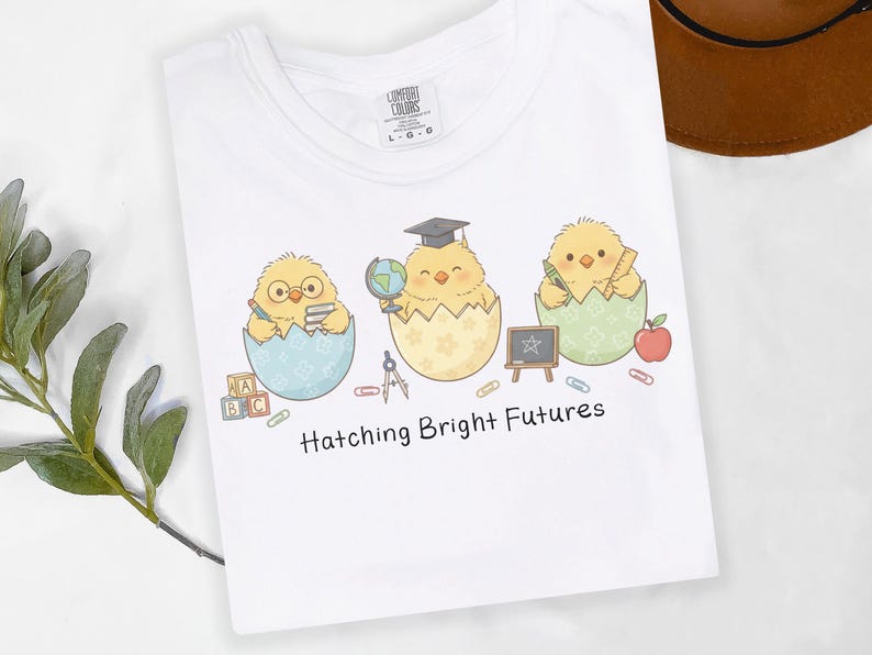 Puede incluir: Camiseta blanca con un dise&ntilde;o de tres pollitos de dibujos animados saliendo de huevos, cada uno con accesorios escolares. El texto "Hatching Bright Futures" est&aacute; impreso debajo. La camiseta tiene una etiqueta Comfort Colors.