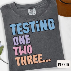 Könnte beinhalten: Dunkelgraues T-Shirt mit dem Schriftzug "TESTING ONE TWO THREE..." in einem farbenfrohen Blockbuchstaben-Design. Das Shirt besteht aus weichem Material und hat einen Rundhalsausschnitt. Das Etikett lautet "Comfort Colors".