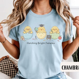Puede incluir: Camiseta azul claro con un dise&ntilde;o de tres pollitos saliendo de huevos, cada uno con &uacute;tiles escolares. El texto "Hatching Bright Futures" est&aacute; impreso debajo. La camiseta es de un material suave.