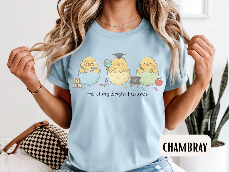 Puede incluir: Camiseta azul claro con un dise&ntilde;o de tres pollitos saliendo de huevos, cada uno con &uacute;tiles escolares. El texto "Hatching Bright Futures" est&aacute; impreso debajo. La camiseta es de un material suave.