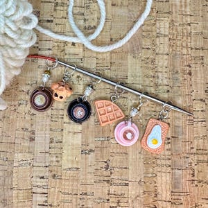 May include: A collection of colorful stitch markers with food-themed charms, including a coffee cup, muffin, waffle, pink coffee, and toast with a fried egg. The markers are attached to a silver knitting needle, perfect for knitters.