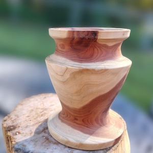 May include: A handcrafted wooden vase in light brown and reddish-brown tones. The vase features a wide base, a narrow neck, and a flared rim. It rests on a wooden stump, with a blurred background of green and grey.