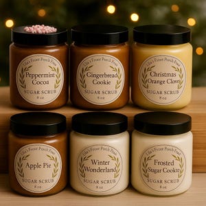 May include: Six jars of sugar scrub with black lids are displayed. Flavors include Peppermint Cocoa, Gingerbread Cookie, Christmas Orange Clove, Apple Pie, Winter Wonderland, and Frosted Sugar Cookie. Each jar has a label with the flavor name and "SUGAR SCRUB 8 oz."