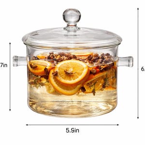 Puede incluir: Una olla de vidrio transparente con tapa, llena de agua, rodajas de naranja y especias. La olla mide 15 cm de ancho y 16 cm de alto, con un asa a cada lado. El contenido parece ser una infusi&oacute;n fragante.
