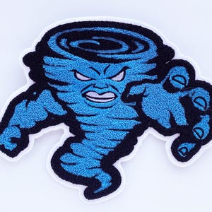 May include: Embroidered patch featuring a blue tornado with an angry face and outstretched arm. The patch has a black outline and a white border. The design is detailed and textured, giving it a three-dimensional appearance. Ideal for jackets, bags, or other fabric items.