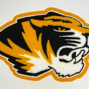 May include: Embroidered tiger head patch with black, gold, and white details. The tiger's face is depicted with sharp teeth and an intense gaze, outlined in gold and black. The patch is likely for apparel or accessories.