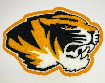 May include: Embroidered tiger head patch with black, gold, and white details. The tiger's face is depicted with sharp teeth and an intense gaze, outlined in gold and black. The patch is likely for apparel or accessories.