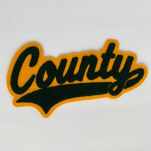 May include: A yellow felt patch with the word "County" in dark green, script lettering. The patch has a dark green banner beneath the word and a yellow border. The patch is on a white background.