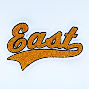 May include: A gold-colored, embroidered patch with the word "East" in a script font. The text is outlined in black and has a white border. The patch has a banner-like design element below the text.