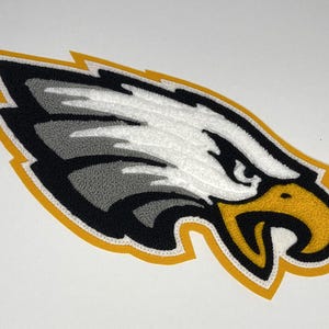 May include: A patch in the shape of an eagle's head, featuring white, gray, black, and yellow colors. The eagle's eye is black, and the beak is yellow. The patch has a yellow border and is likely for apparel or accessories.