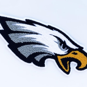 May include: Embroidered patch featuring an eagle head design. The eagle has a white head, gray and black wing, and a gold beak. The patch is outlined in white and is likely for apparel or accessories.