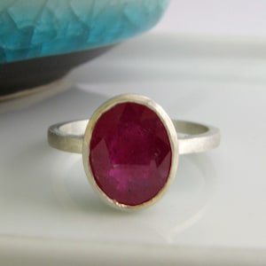 K&ouml;nnte beinhalten: Ein silberner Ring mit einem gro&szlig;en, ovalen Rubin-Edelstein. Der Rubin ist tiefrot und hat einen facettierten Schliff.