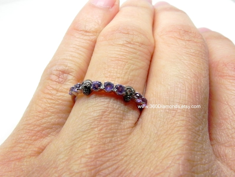 May include: A silver band ring with a skull design and purple gemstones. The ring is on a person's finger.