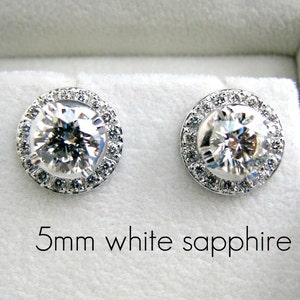 May include: A pair of silver earrings with a 5mm white sapphire center stone surrounded by a halo of smaller diamonds.