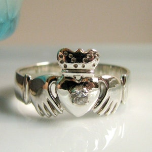 Peut inclure: Une bague Claddagh en argent avec un cœur, des mains et une couronne. Le cœur a une petite pierre claire au centre.