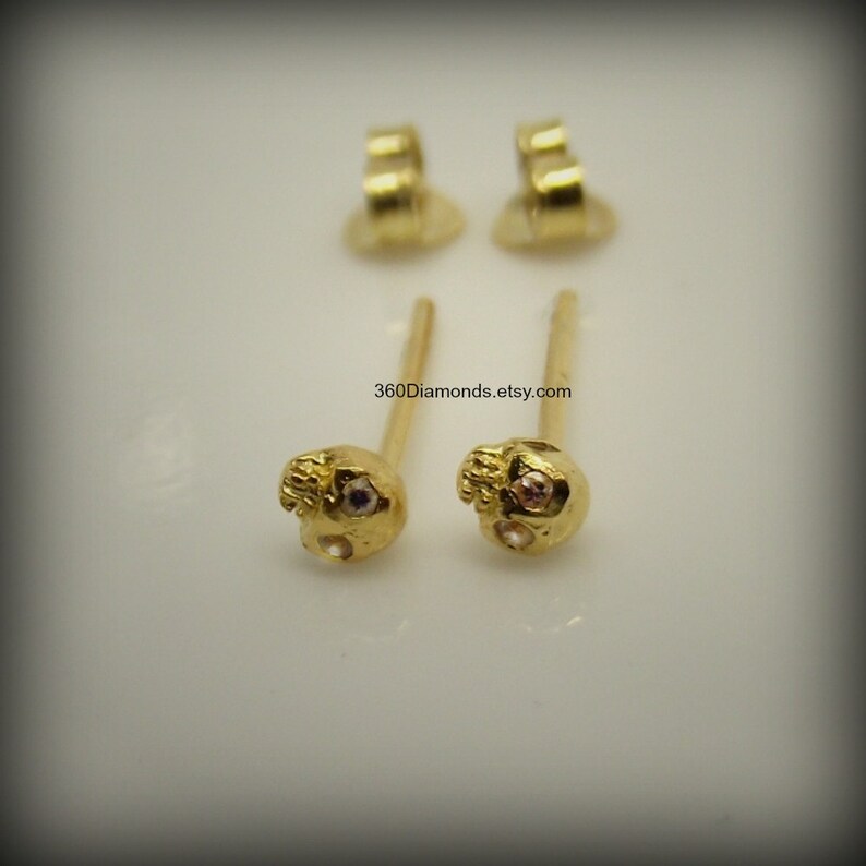 May include: A pair of gold skull earrings with small clear and pink gemstones for the eyes. The earrings have a post and butterfly back.
