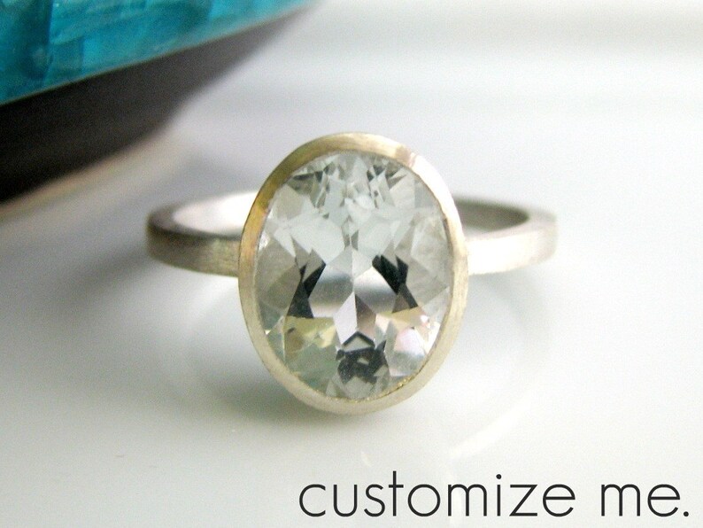 K&ouml;nnte beinhalten: Ein silberner Ring mit einem gro&szlig;en, ovalen, klaren Edelstein. Der Ring ist in einer einfachen Zargenfassung gefasst. Der Edelstein ist facettiert und hat einen leichten Blaustich. Der Ring befindet sich auf einem wei&szlig;en Hintergrund. Personalisieren Sie mich.