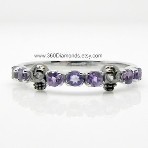 May include: A silver band ring with a skull design and purple gemstones. The skull has a single diamond for an eye.