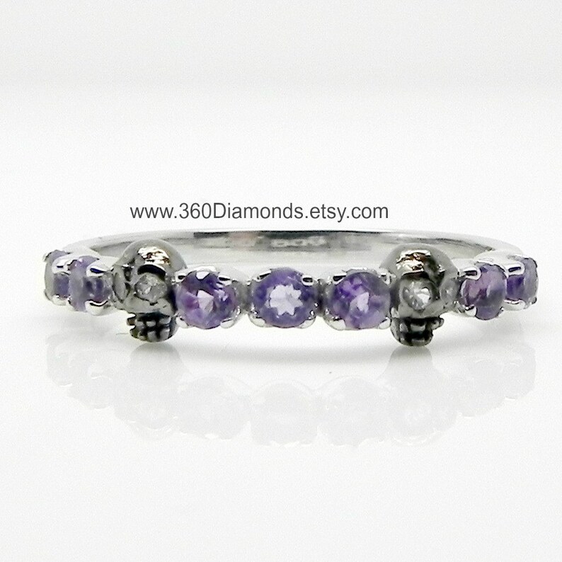 May include: A silver band ring with a skull design and purple gemstones. The skull has a single diamond for an eye.