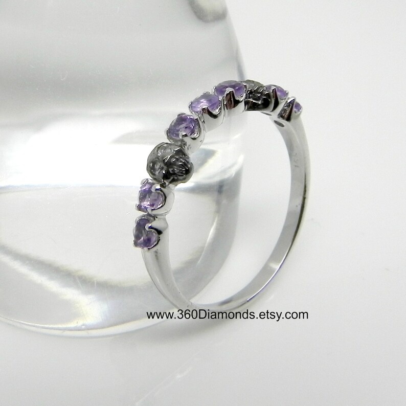 May include: A silver band ring with a row of small, light purple amethyst gemstones set in a curved, prong setting. The ring is on a white background.
