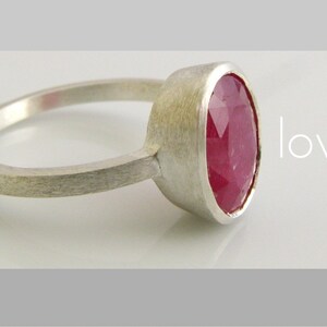 K&ouml;nnte beinhalten: Ein silberner Ring mit einem gro&szlig;en, ovalen, facettierten Rubin-Edelstein. Der Ring ist schlicht und elegant, mit einer geb&uuml;rsteten Silber-Oberfl&auml;che. Der Text "love." ist im Hintergrund sichtbar.