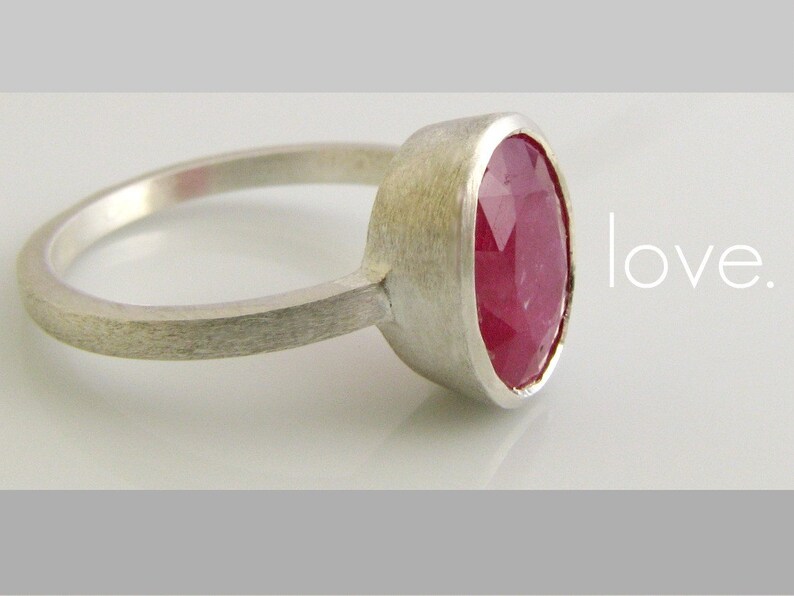 K&ouml;nnte beinhalten: Ein silberner Ring mit einem gro&szlig;en, ovalen, facettierten Rubin-Edelstein. Der Ring ist schlicht und elegant, mit einer geb&uuml;rsteten Silber-Oberfl&auml;che. Der Text "love." ist im Hintergrund sichtbar.