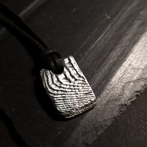 May include: A silver pendant necklace with a textured, rectangular design. The pendant is attached to a black cord.
