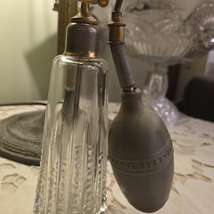 May include: An antique perfume bottle with a clear, textured glass body and a gold-colored metal top and sprayer. The bulb-shaped atomizer is gray and has embossed text. The bottle is displayed on a white lace tablecloth.
