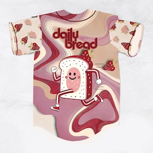 Op de afbeelding: Een honkbalshirt met een cartoon brood karakter dat over de voorkant rent. Het shirt heeft een roze en crème kleurenschema met de woorden "daily bread" in kastanjebruin. De mouwen hebben een aardbeienpatroon.