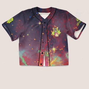 Puede incluir: Camiseta de béisbol con un estampado de galaxia vibrante en tonos morados, rojos y amarillos. La camiseta presenta un gráfico de un extraterrestre de dibujos animados y la palabra "SUBTLE" en amarillo. Ribete negro en el cuello, botones y mangas.