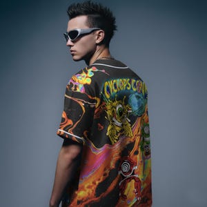 May include: Black baseball jersey with a vibrant, psychedelic design featuring a cartoon cyclops and the text "CYCLOPS CO." The jersey has white trim and short sleeves. The background is a gradient of orange, yellow, and blue.