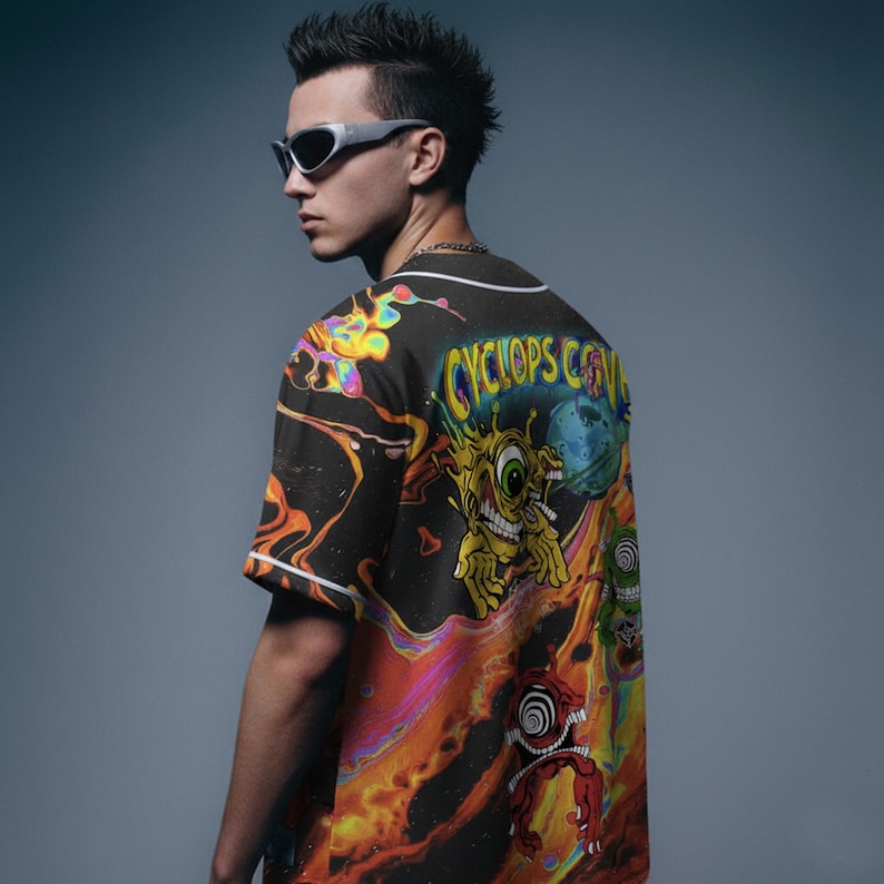 May include: Black baseball jersey with a vibrant, psychedelic design featuring a cartoon cyclops and the text "CYCLOPS CO." The jersey has white trim and short sleeves. The background is a gradient of orange, yellow, and blue.