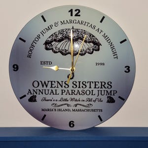 May include: A circular clock with a pale blue face, black text, and a black parasol illustration. The clock hands are gold. Text includes "ROOFTOP JUMP & MARGARITAS AT MIDNIGHT" and "OWENS SISTERS ANNUAL PARASOL JUMP".