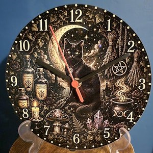 May include: A round wall clock featuring a black cat sitting before a crescent moon and starry sky. The clock face displays numbers 1 through 12, glass bottles labelled Wormwood and Nightshade, a steaming cauldron, crystals, and mushrooms.