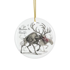 以下が含まれることがあります： 金色の紐が付いた白いセラミック製の丸いクリスマスオーナメント。 「Christmas Is Coming...」のテキストが入った、骸骨のトナカイのイラストが特徴です。 トナカイはグレーで、大きな角があります。