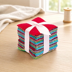 May include: A stack of square fabric swatches featuring a variety of vibrant colors including red, blue, green, and yellow. The fabric bundle is tied together with a light pink grosgrain ribbon, forming a neat, compact square-shaped presentation.