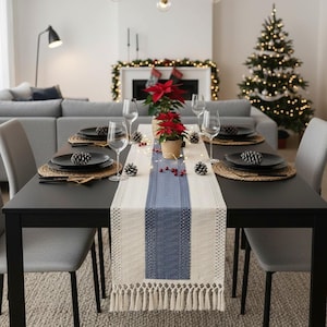 以下が含まれることがあります： お祝いの食事のためにセットされたダイニングテーブル。黒いテーブルには、黒いお皿、ワイングラス、青いストライプの織りランナーがセットされています。ポインセチアの植物と松ぼっくりがテーブルを飾っています。背景にはクリスマスツリーがあります。