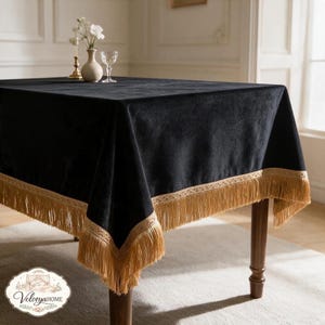 Op de afbeelding: Een vierkant zwart fluwelen tafelkleed met een decoratieve gouden franje langs de randen. De stof heeft een zachte, matte textuur en de gouden rand bevat een geweven patroon boven de hangende kwastjes die over de hoeken van de tafel vallen.