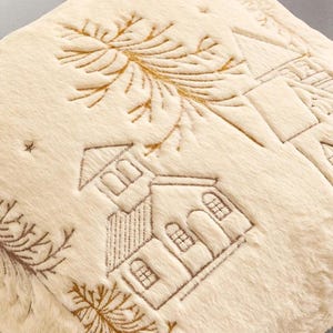 Pu&ograve; includere: Tessuto color crema e felpato con scena invernale ricamata. Presenta case, alberi e stelle cucite in marrone. La consistenza morbida e il ricamo dettagliato creano un'estetica accogliente e stagionale.