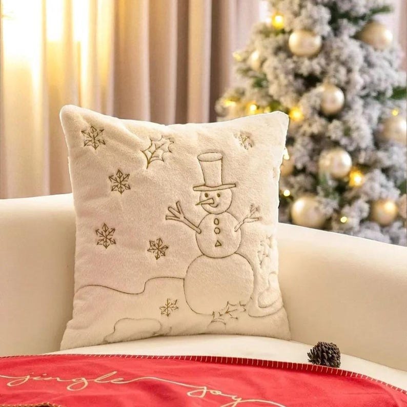 Pu&ograve; includere: Cuscino decorativo color crema con un design a pupazzo di neve. Il cuscino presenta un pupazzo di neve ricamato con un cappello a cilindro, circondato da fiocchi di neve. Il cuscino &egrave; su un divano bianco, con un runner da tavolo rosso in primo piano e un albero di Natale sullo sfondo.