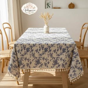 Könnte beinhalten: Ein rechteckiges beiges Tischtuch mit blauem Blumenmuster und weißen Quasten. Das Tischtuch liegt auf einem Holztisch mit zwei Holzstühlen auf beiden Seiten. Eine weiße Vase mit Weizen steht auf dem Tisch.