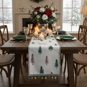 Op de afbeelding: Een feestelijke eettafel gedekt voor een feestmaaltijd. De houten tafel is versierd met een witte loper met een hert- en boompatroon. Groene borden, gouden onderborden en elegant glaswerk completeren de setting, met een bloemstuk en brandende kaarsen.